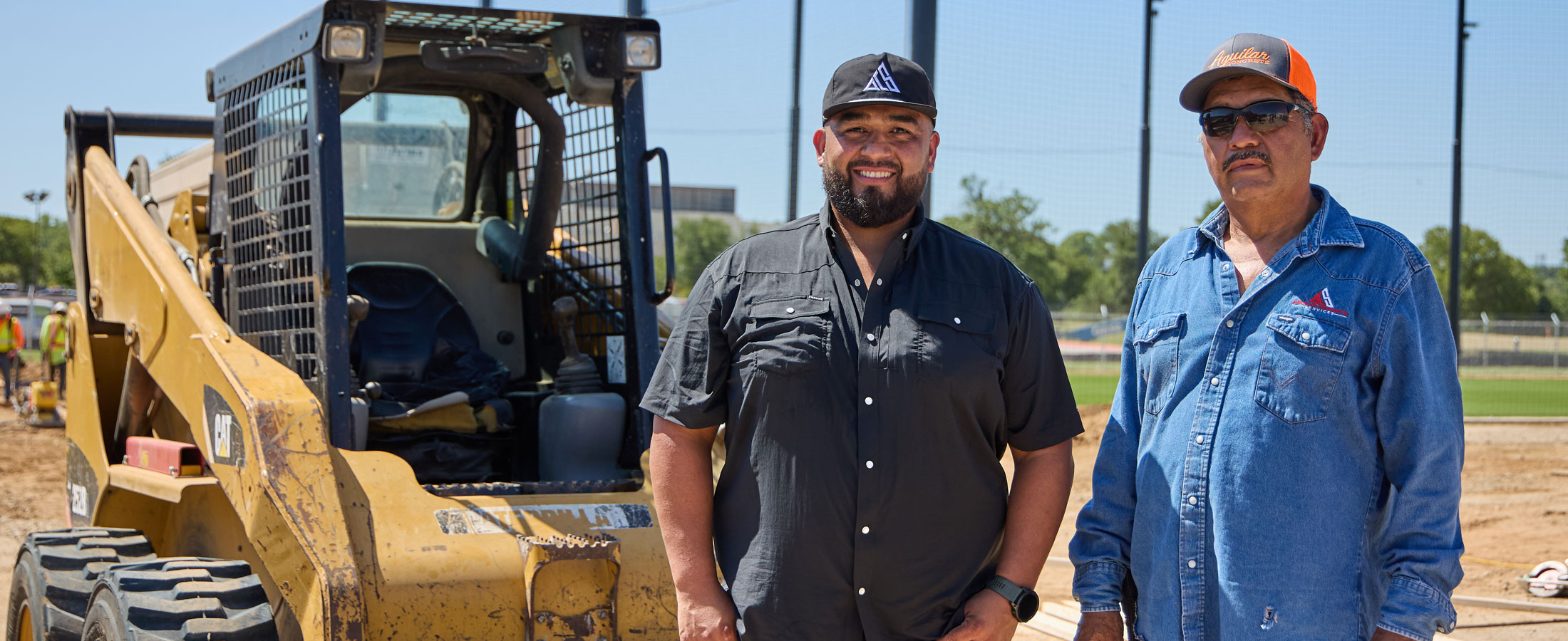 Two men, Benny and Leno Aguilar, stand side by side at a construction site in front of heavy machinery.